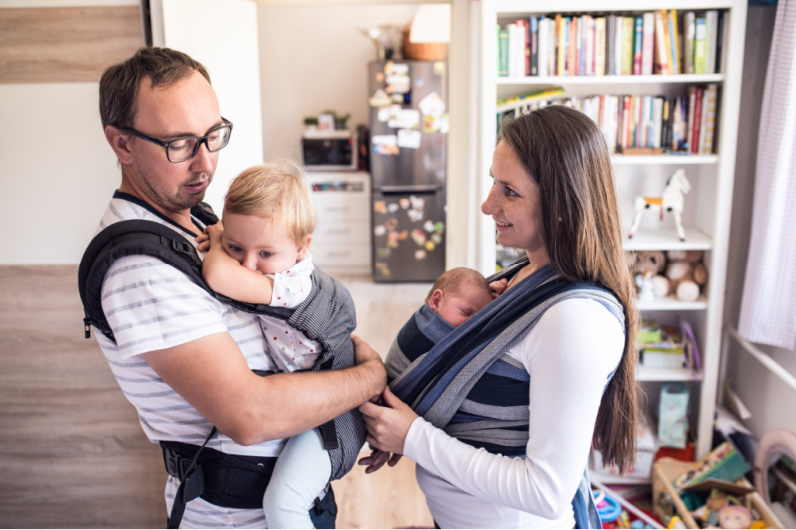two parents with babies in baby carriers, heavy bookshelf in background