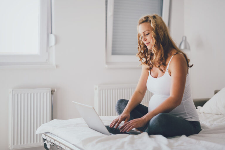 pregnant woman sitting on a bed with a laptop adding items to her baby registry