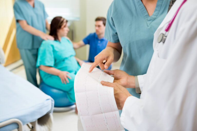 mom trying to have natural birth in hospital, sitting on birthing ball with husband and nurse, doctors examining charts