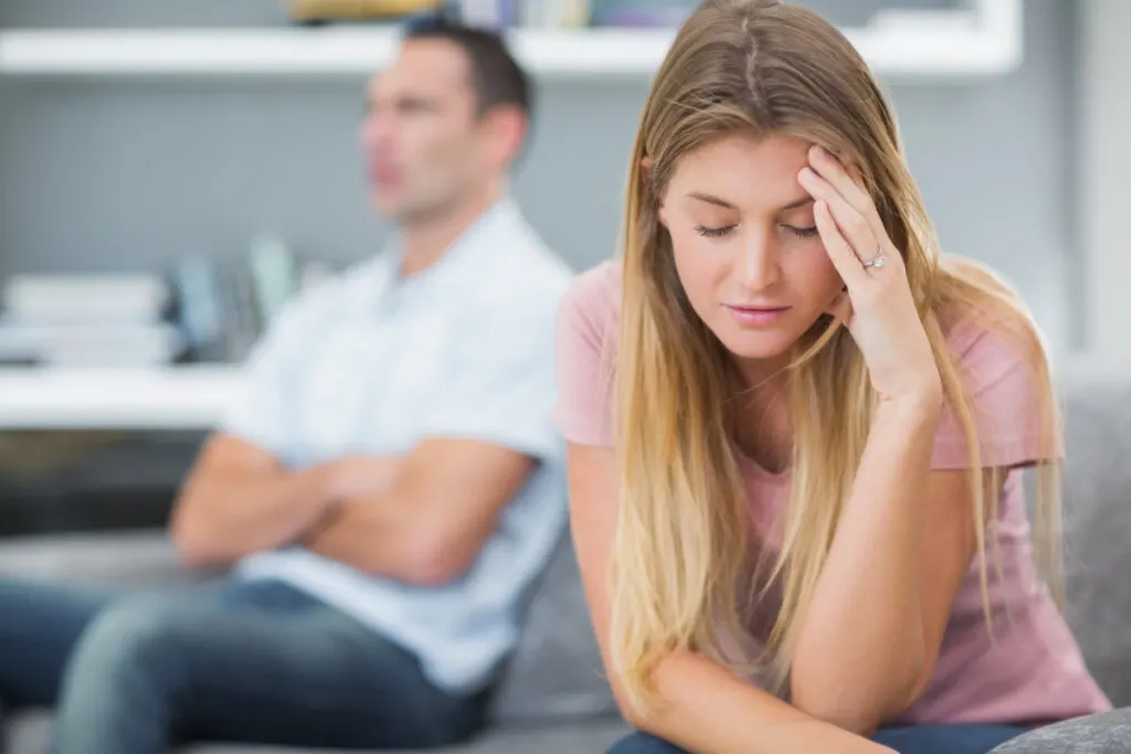 married couple unhappy in marriage after baby, wife holding hand to forehead, husband crossed arms in background