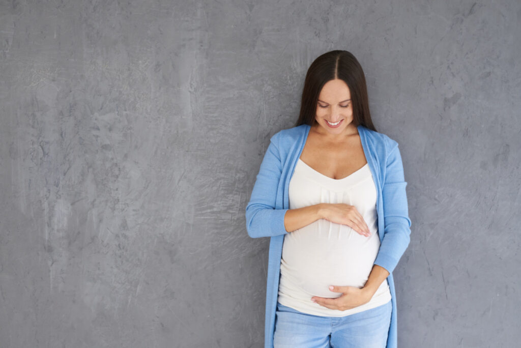 pregnant woman standing in front of gray wall, hands framing pregnant belly, praying for her unborn baby