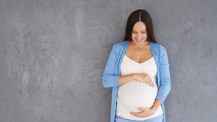 pregnant woman standing in front of gray wall, hands framing pregnant belly, praying for her unborn baby