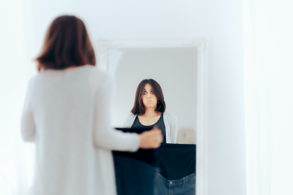pregnant woman standing in front of mirror holding up maternity jeans, feeling unattractive