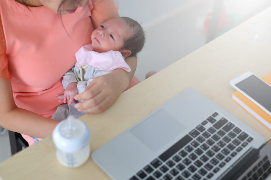 work from home mom holding a newborn while trying to work, sitting at desk