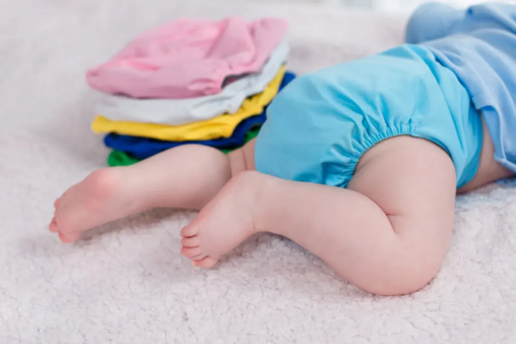 The child lies near a stack of multi-colored cloth diaper covers on a changing table