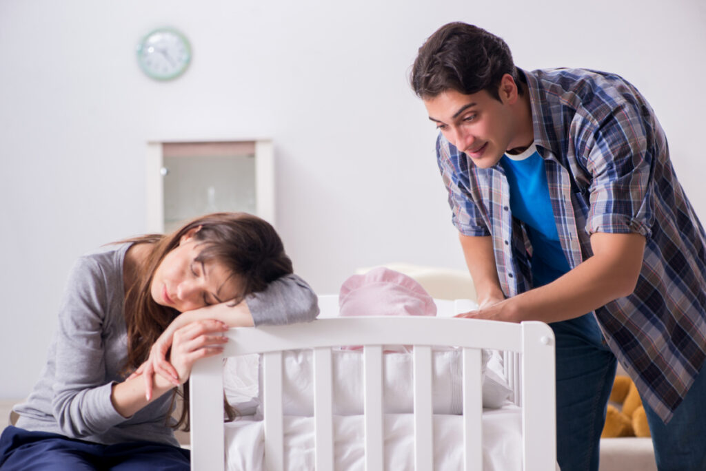 tired new mom resting her head on crib while dad takes care of baby