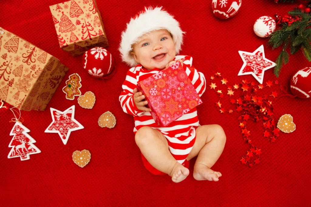 baby with Santa hat holding Christmas present surrounded by ornaments on red background - baby's first Christmas theme