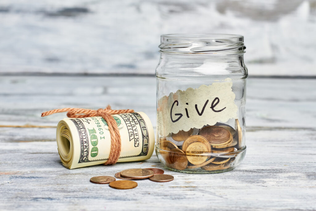glass jar labeled "give" with coins inside and 100 dollar bill sitting next to it, sinking fund for gifts concept