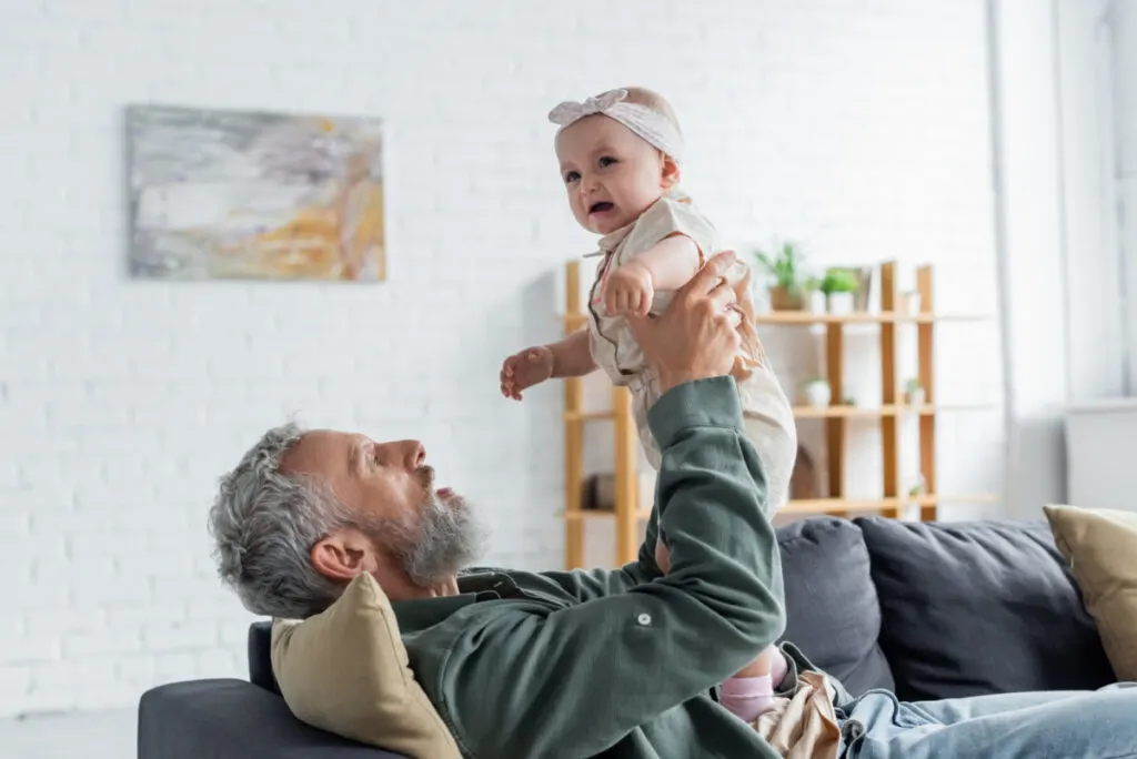 grandfather sitting on couch holding not happy infant girl up in the air