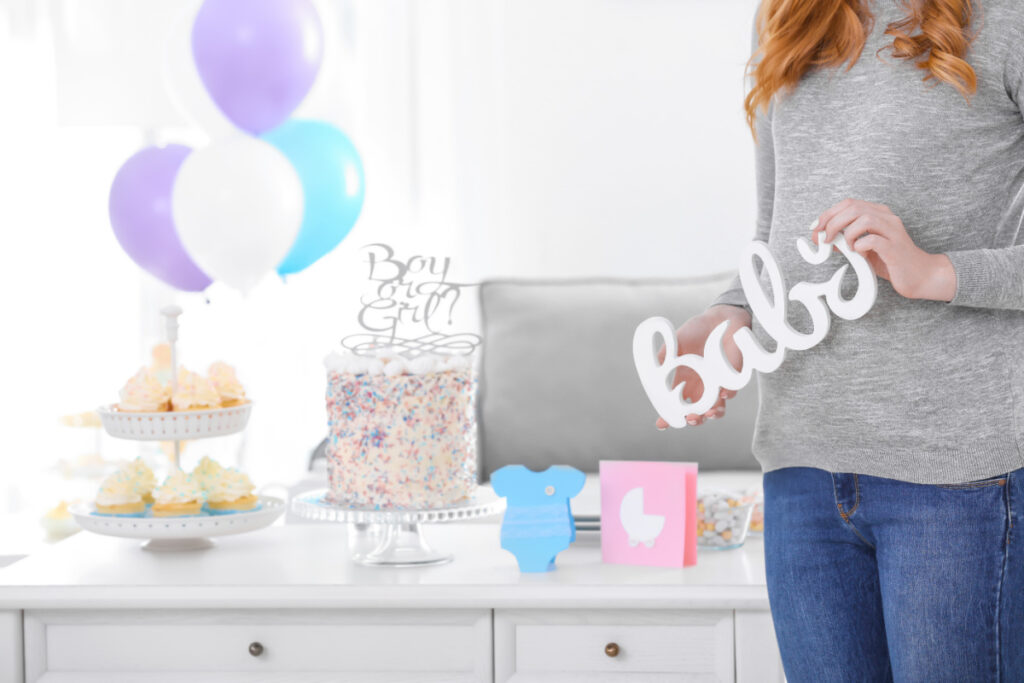 baby shower dessert and game prize display table, with pregnant woman holding a white, wooden "baby" sign in front of her belly