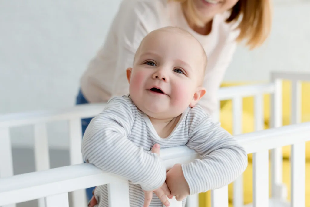 baby putting arms over crib bars with mom standing behind on the opposite side of the crib