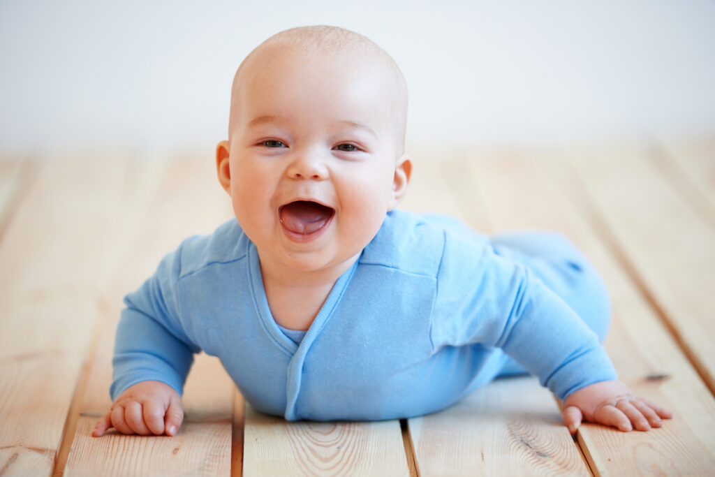 smiling baby boy dressed in blue pajamas, laying on belly on the floor, pushing up with arms.