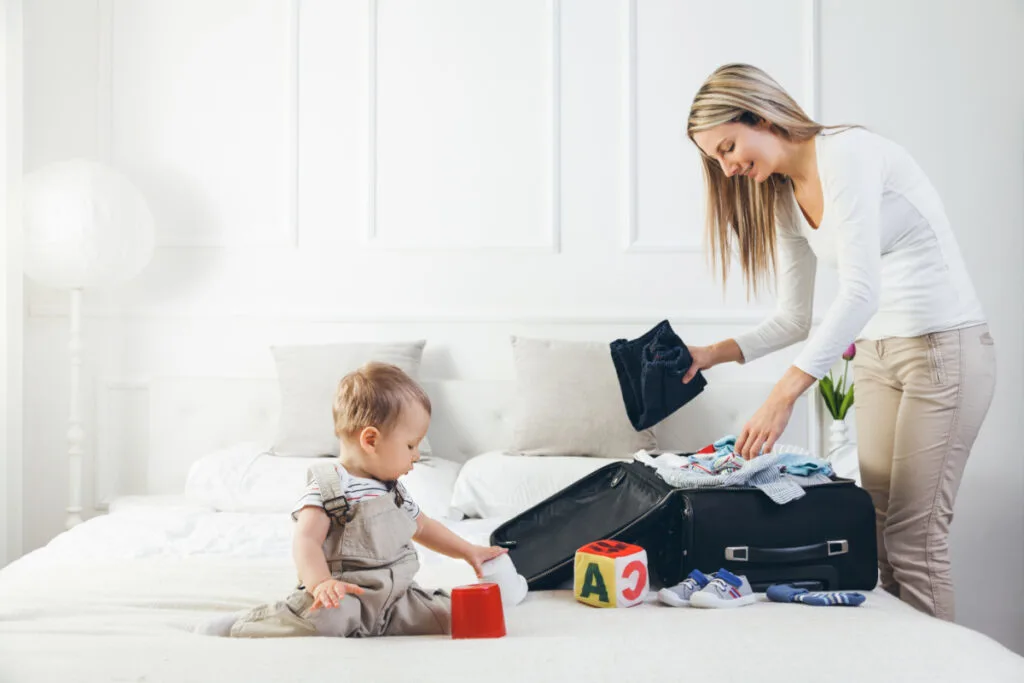 mom packing infant travel essentials in a suitcase while baby plays on bed.