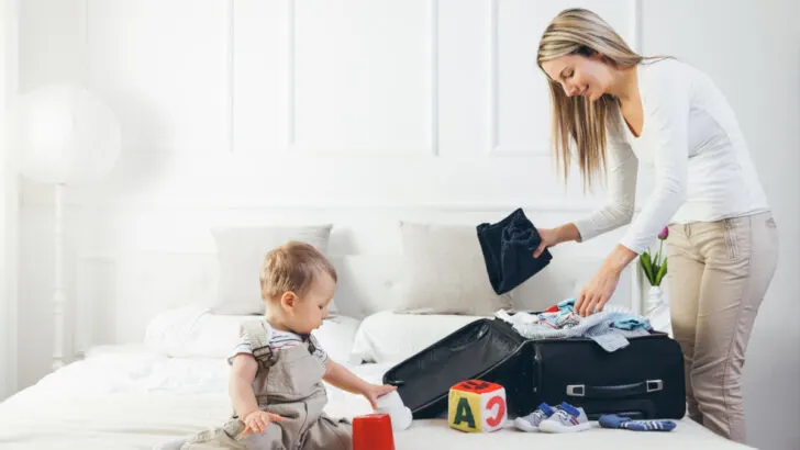 mom packing infant travel essentials in a suitcase while baby plays on bed.
