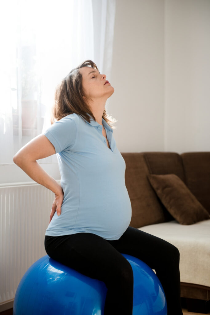 Pregnant woman sitting on fit ball trying to relieve backache at home.