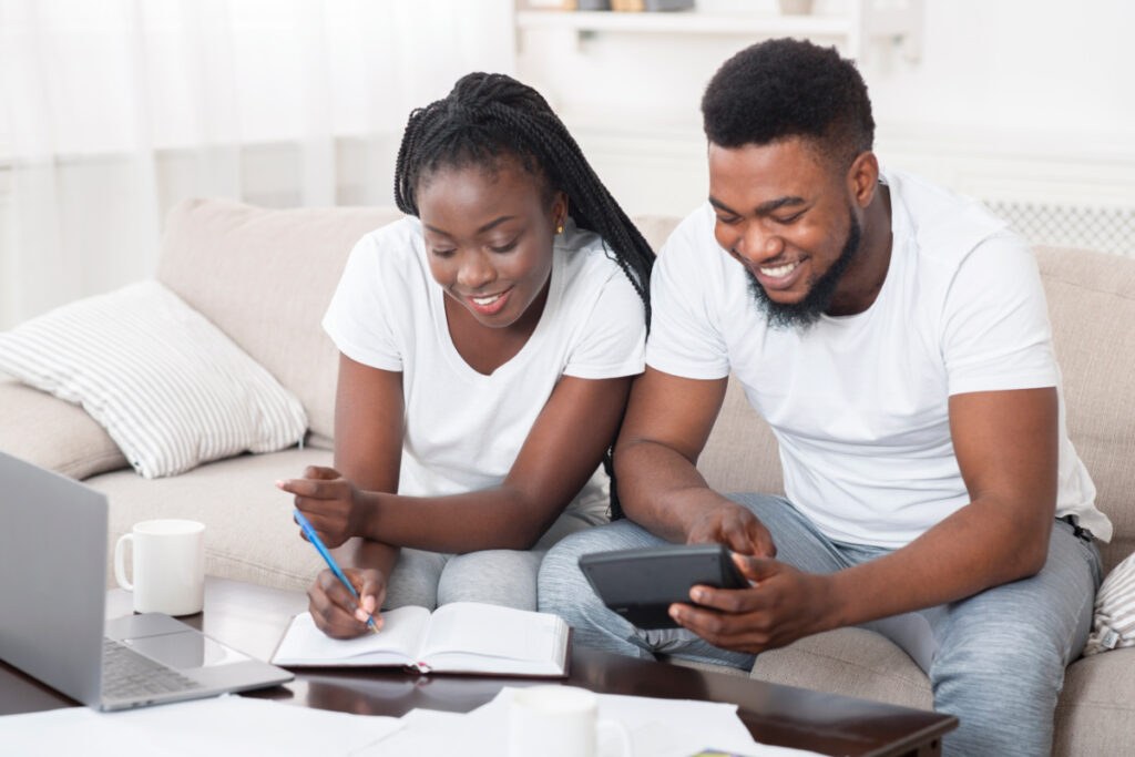 smiling black couple sitting on a couch with a notebook and calculator, making a budget together.