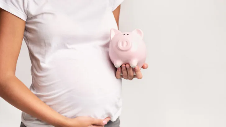 pregnant woman in white t-shirt holding her belly and a pink piggy bank, thinking about having a baby on a tight budget.