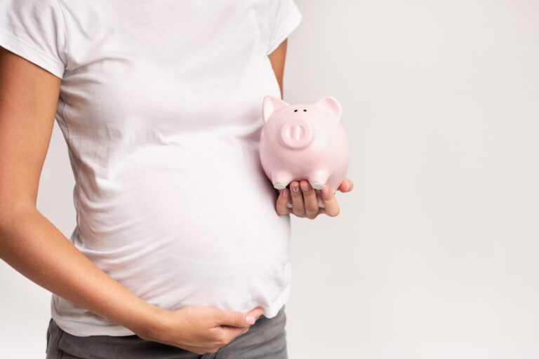 pregnant woman in white t-shirt holding her belly and a pink piggy bank, thinking about having a baby on a tight budget.
