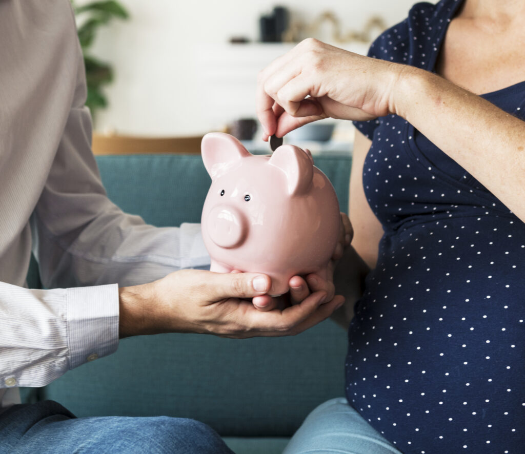 pregnant woman and partner sitting on couch, holding piggy bank between them, saving money for a baby.