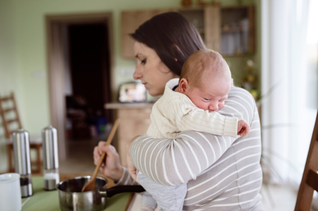 newly postpartum mom holding her newborn while trying to cook a meal.
