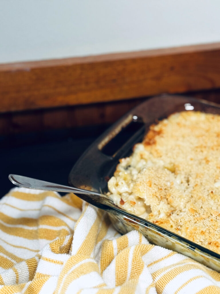 baked Mac and cheese in glass pyrex on counter with serving spoon and yellow striped dish towel.