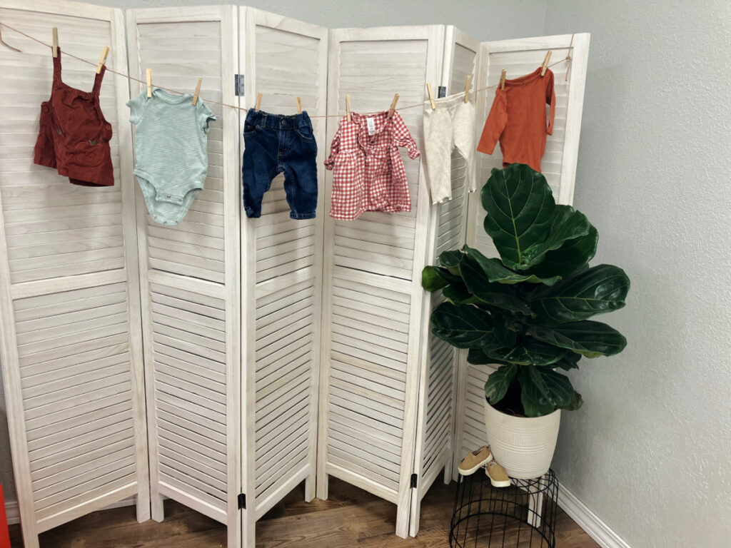 baby clothes strung up on white accordion-style room divider, fiddle leaf fig tree on stand with baby shoes.