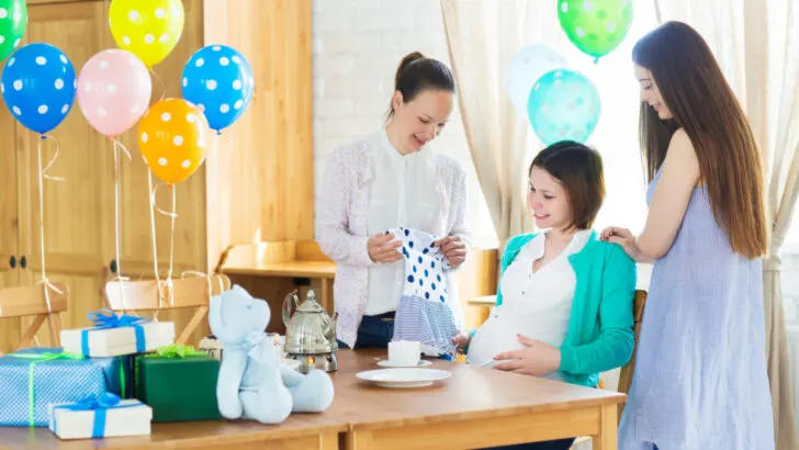 pregnant woman sitting at table at a baby shower, women showing her a onesie that's part of a baby shower game.