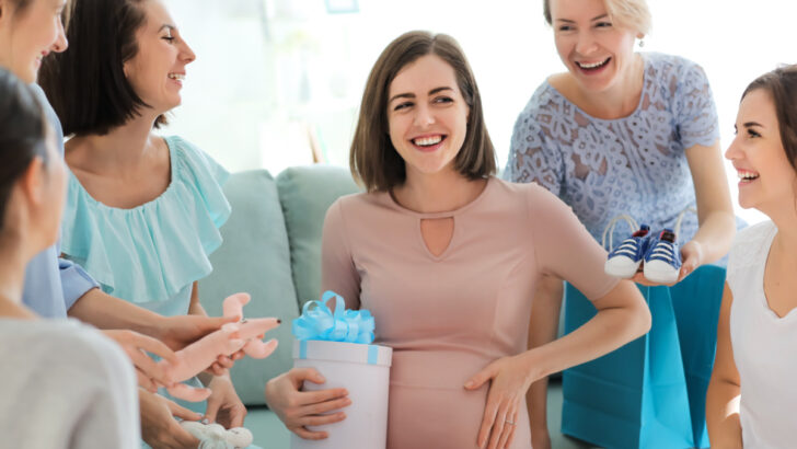 pregnant woman at baby shower holding gift next to pregnant belly and smiling, surrounded by happy guests.