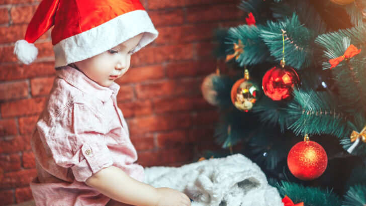 1-year-old with Santa hat on, sitting by Christmas tree looking at gift.