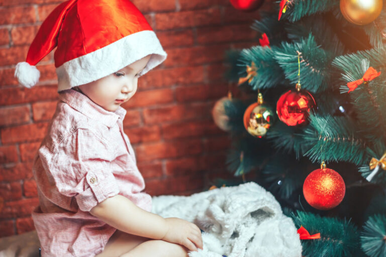 1-year-old with Santa hat on, sitting by Christmas tree looking at gift.
