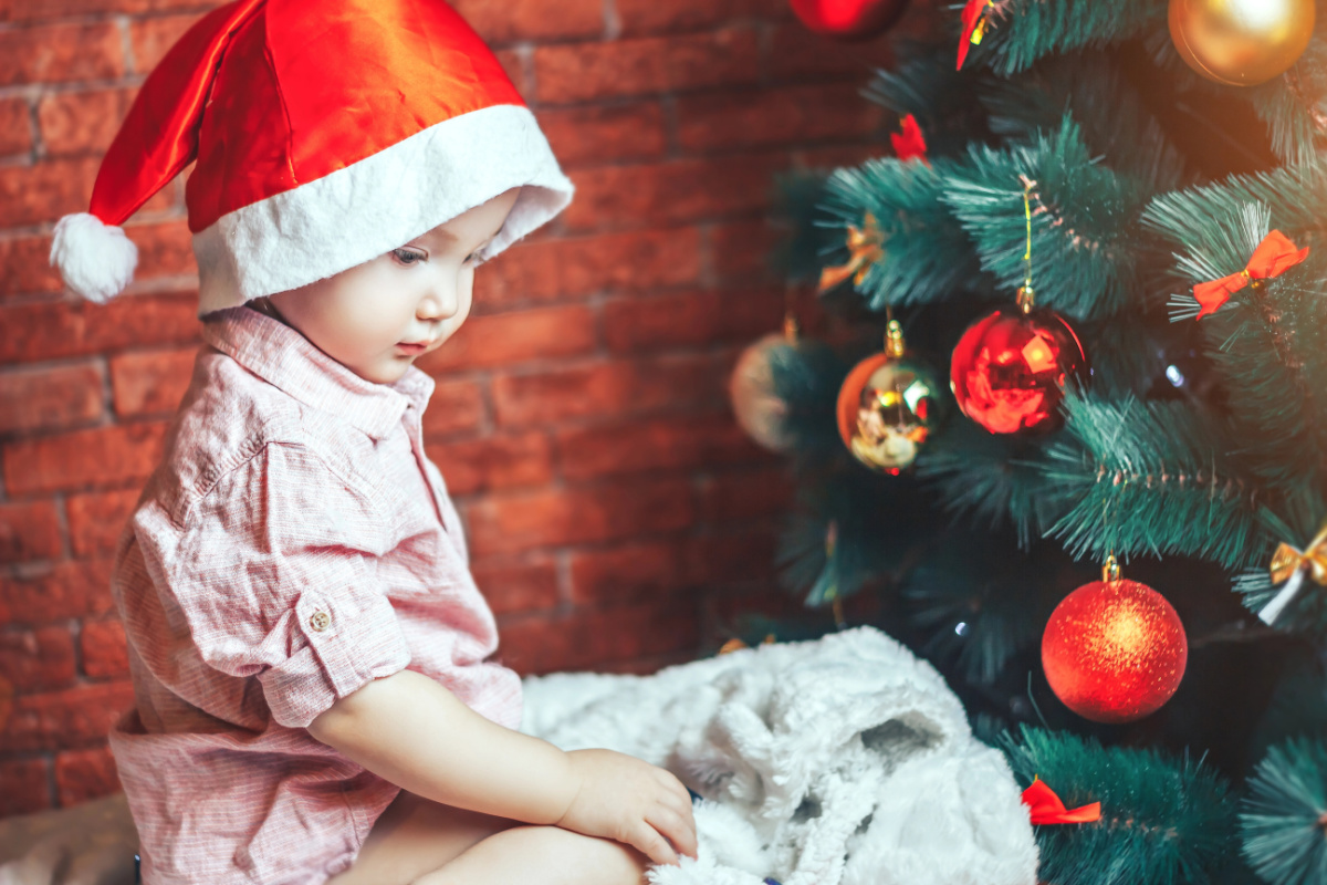 1-year-old with Santa hat on, sitting by Christmas tree looking at gift.