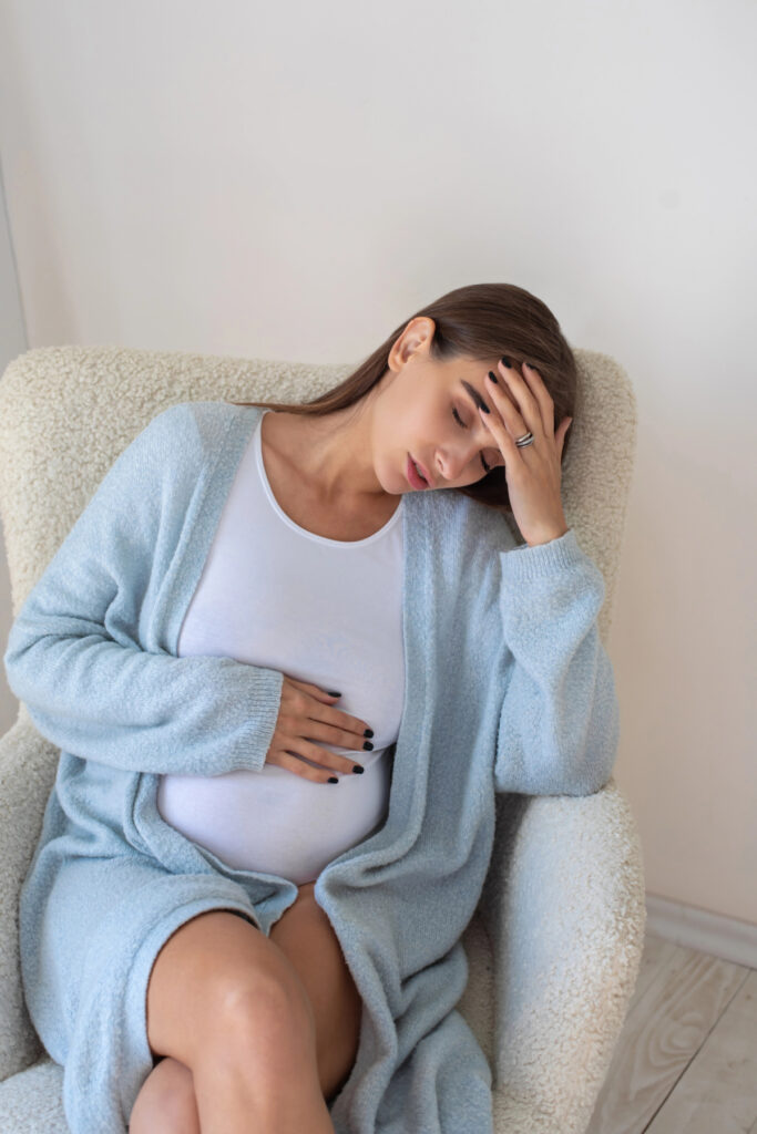tired pregnant woman sitting up in chair with eyes closed and hand on forehead.