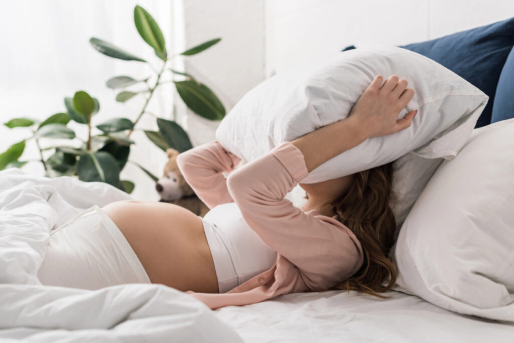 woman struggling with pregnancy insomnia, pillow over face unable to sleep.