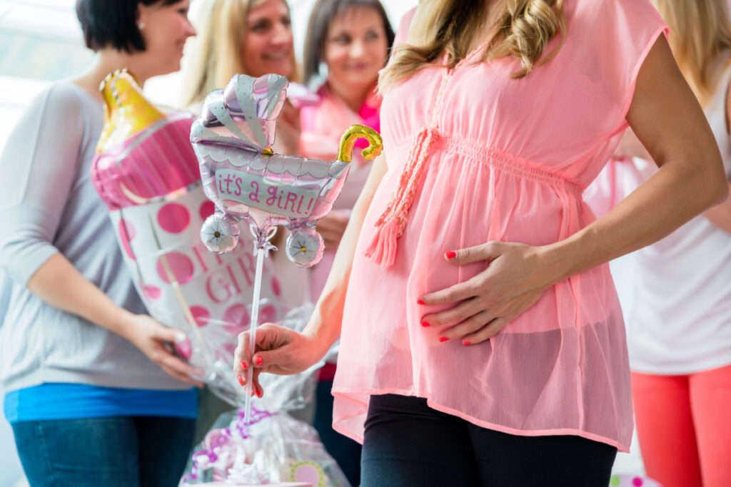 mom to be at baby shower, holding "It's a girl" balloon with hand on pregnant belly.
