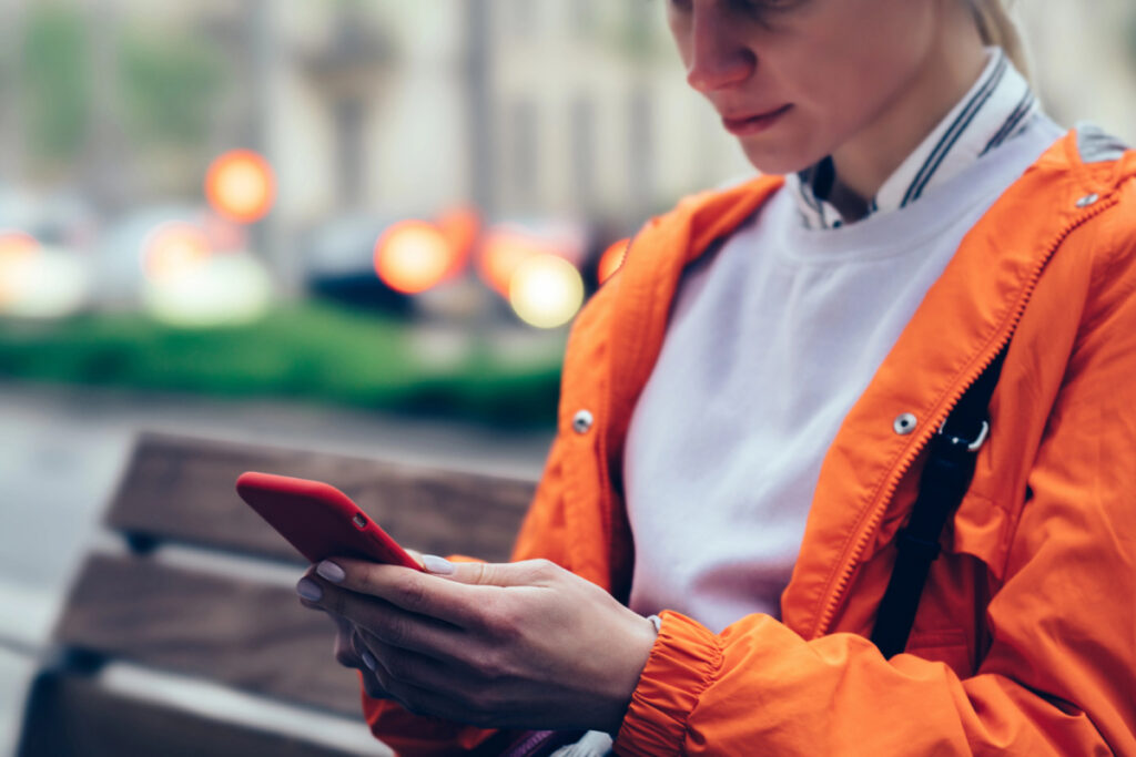 woman in orange jacket sitting on park bench, texting new parents of their arrival for a newborn visit.