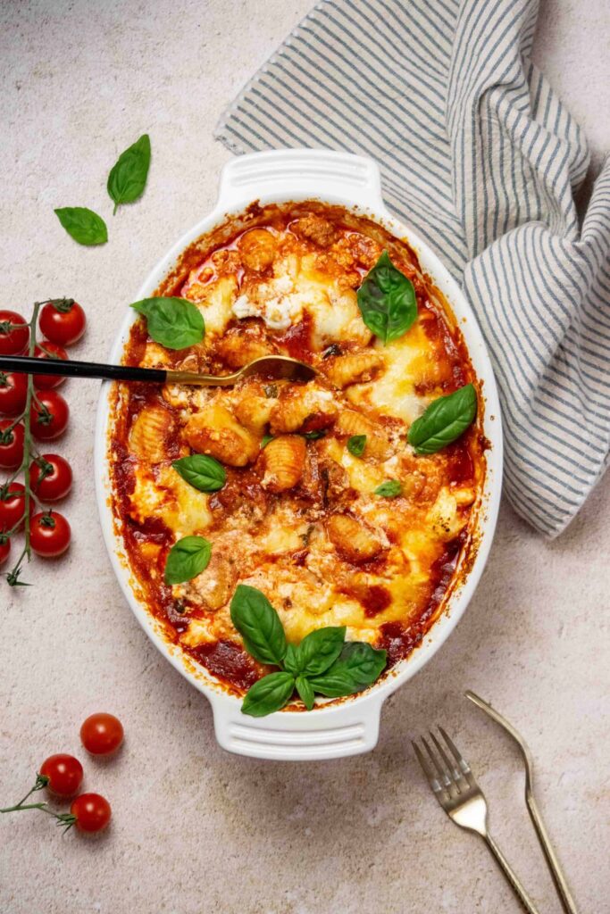 three cheese baked gnocchi casserole in white casserole dish with blue and white striped dish towel and grape tomatoes nearby.