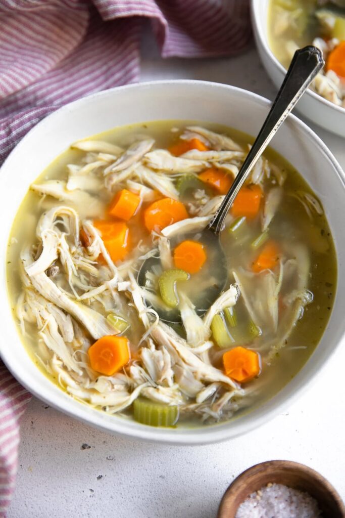 chicken soup without noodles in white bowl with striped dish towel in background.