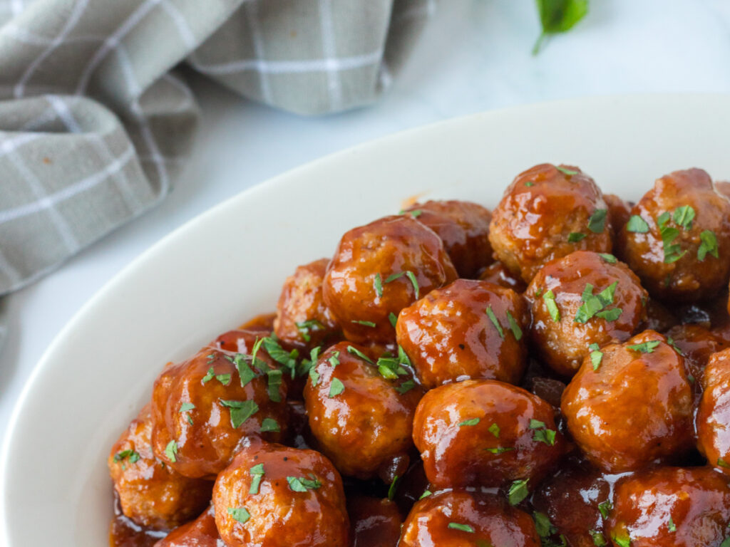 cranberry bbq meatballs on white serving platter with light gray checked dishtowel in background.