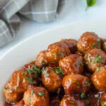 cranberry bbq meatballs on white serving platter with light gray checked dishtowel in background.