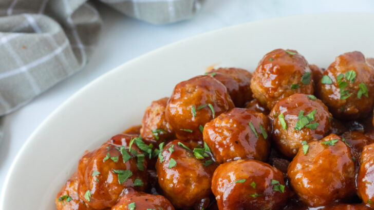 cranberry bbq meatballs on white serving platter with light gray checked dishtowel in background.