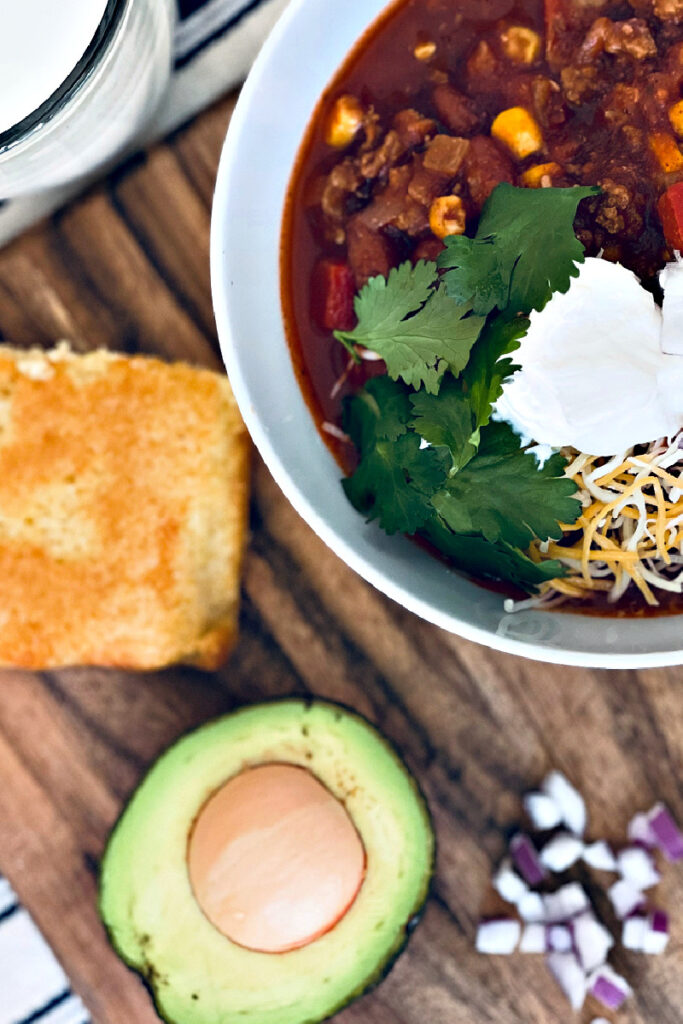 stovetop turkey chili in a bowl with sour cream, shredded cheese and cilantro, on cutting board with cornbread and avocado half.