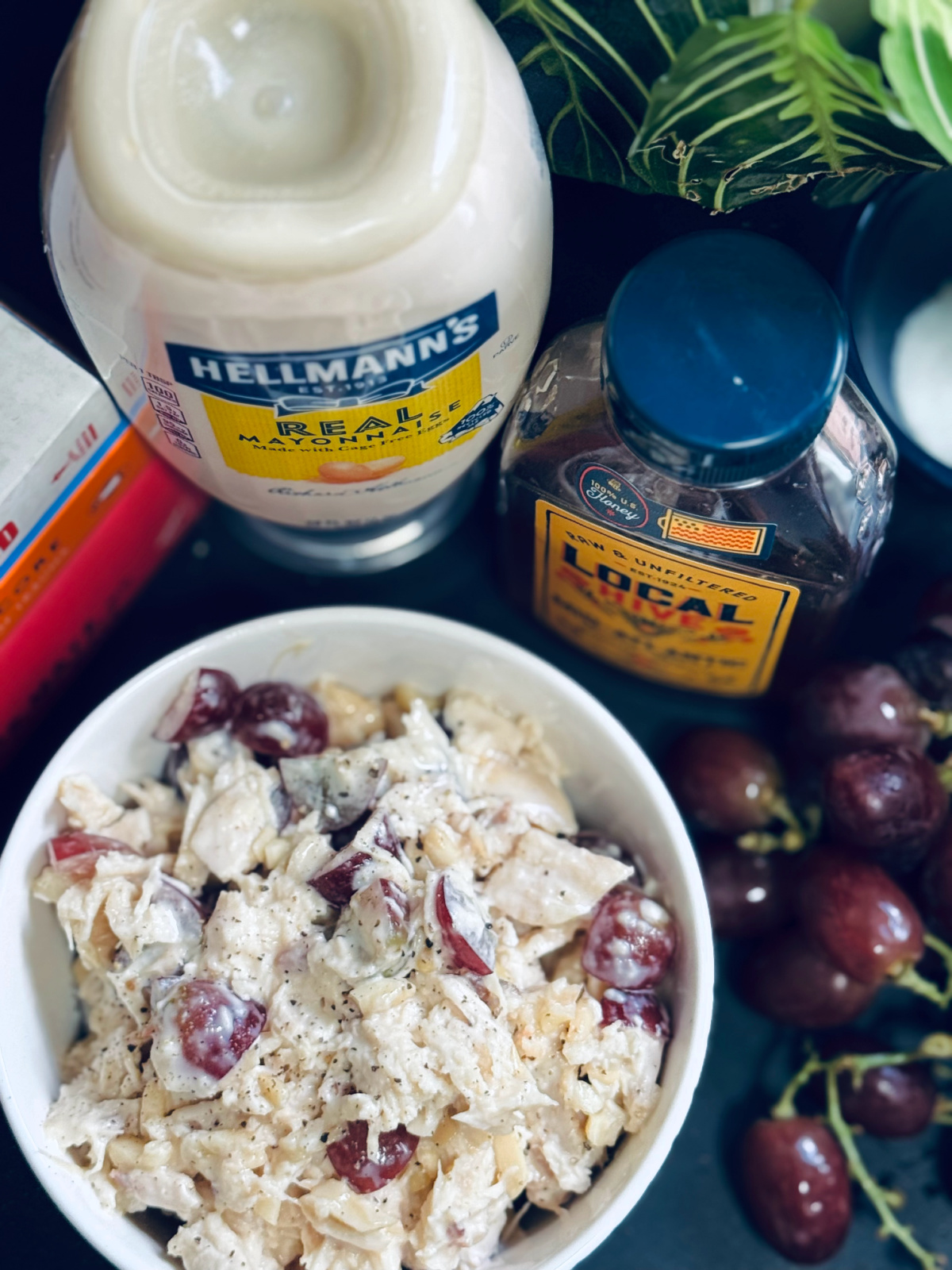 chicken salad in white bowl with mayonnaise, honey and grapes nearby.