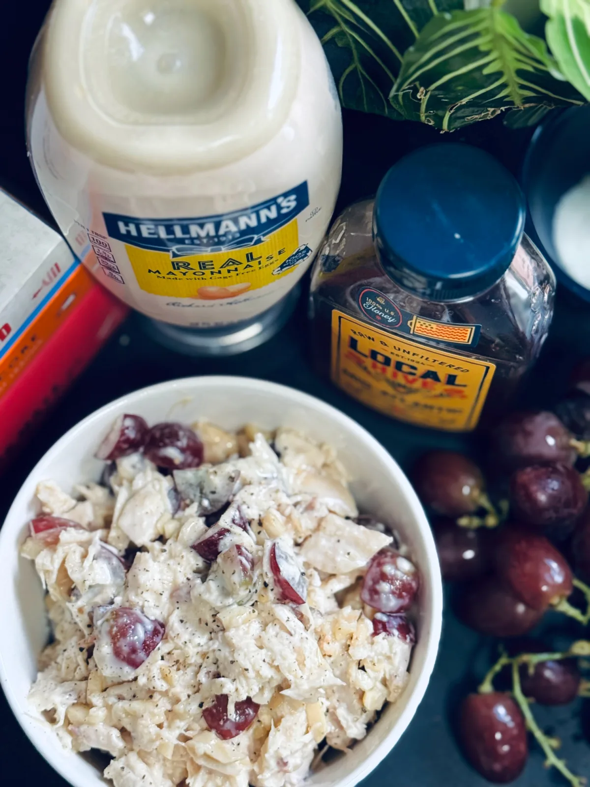 chicken salad in white bowl with mayonnaise, honey and grapes nearby.
