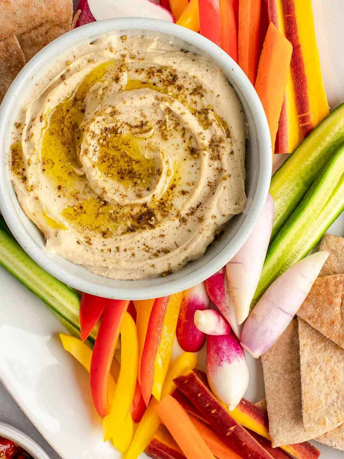 homemade hummus in a white bowl surrounded by vegetables and pita chips.