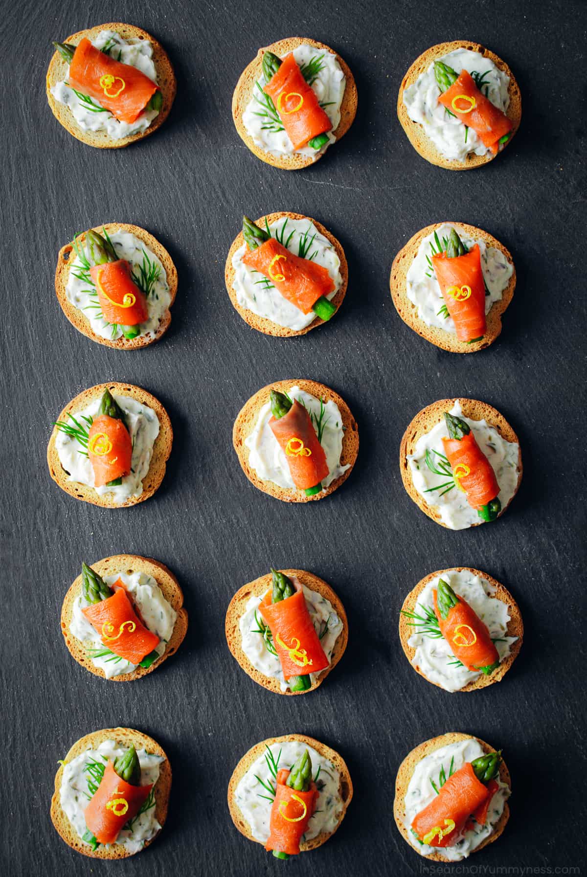 salmon crostini appetizers assembled and lined up on black countertop.